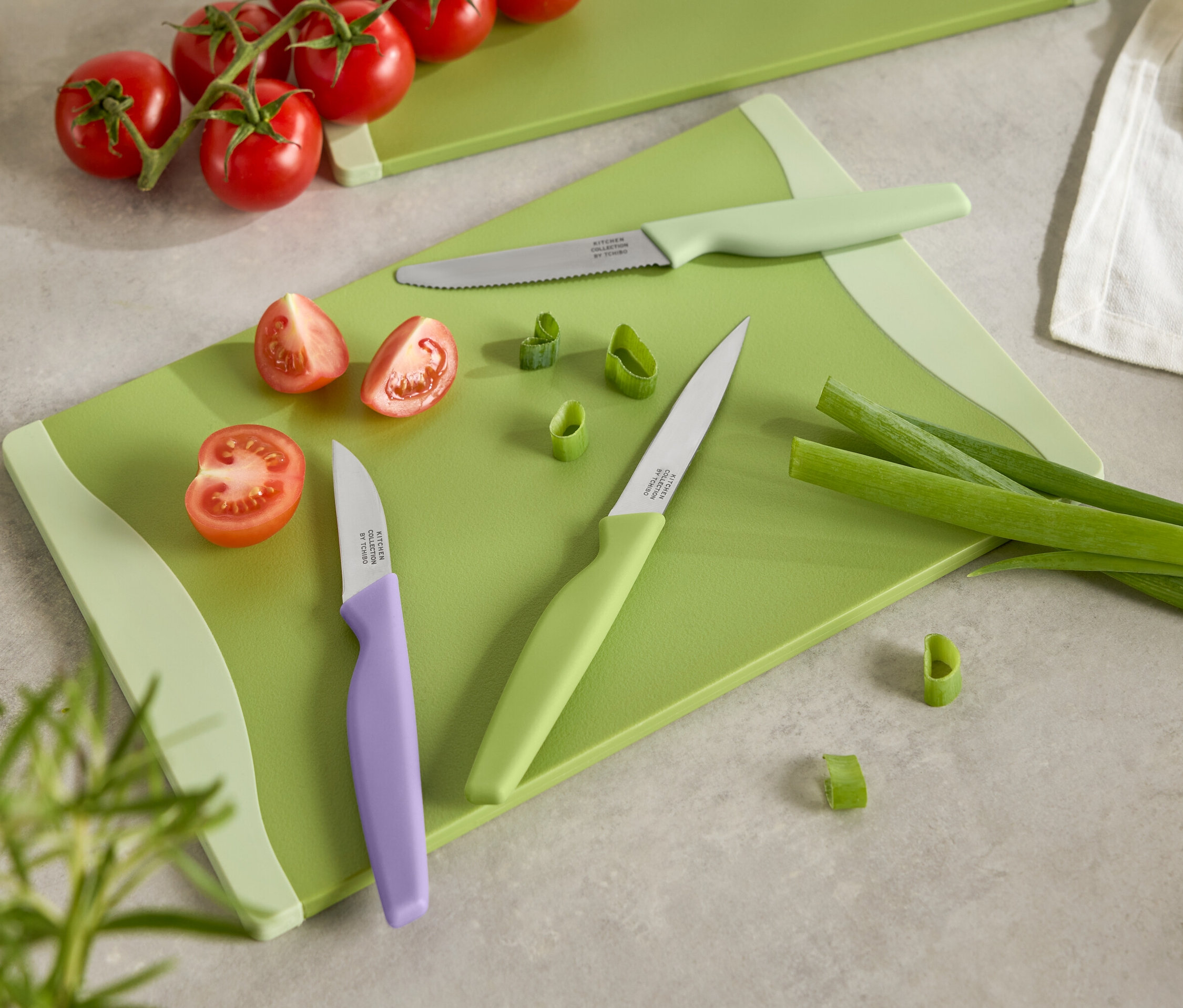 Planche à découper verte avec tomates coupées, oignons nouveaux et trois couteaux à manches colorés.