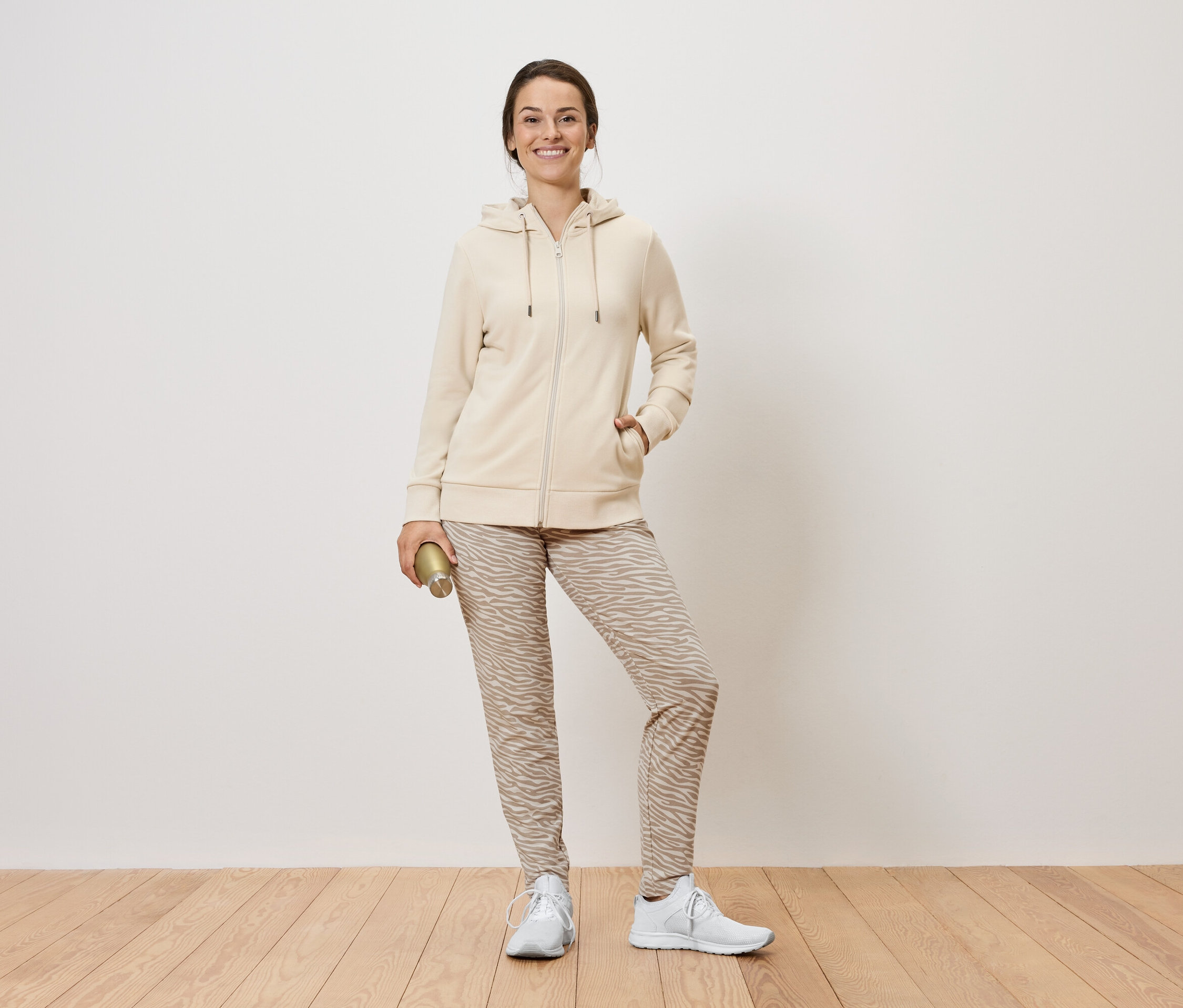 Une femme debout tenant une bouteille dorée. Elle porte un sweat à capuche zippé beige, un pantalon à motifs et des baskets blanches.