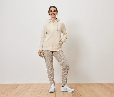 Une femme debout tenant une bouteille dorée. Elle porte un sweat à capuche zippé beige, un pantalon à motifs et des baskets blanches.