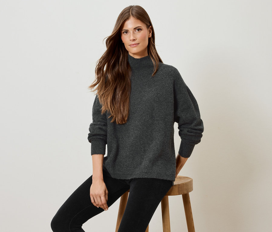 Une femme est assise sur un tabouret en bois, portant un pull gris et un pantalon noir.