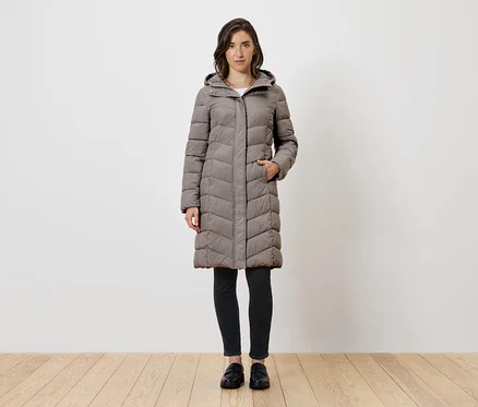 Une femme debout portant un long manteau gris en duvet avec capuche, des leggings noirs et des chaussures noires.
