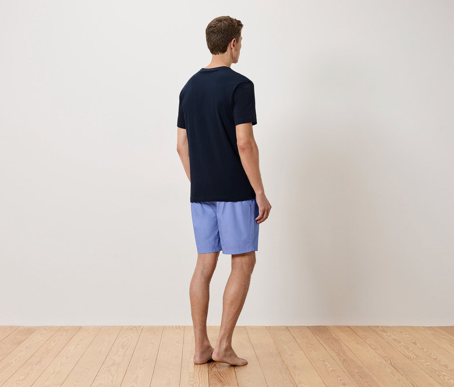Un homme se tient dos à la caméra, il porte un t-shirt bleu marine et un short bleu clair.