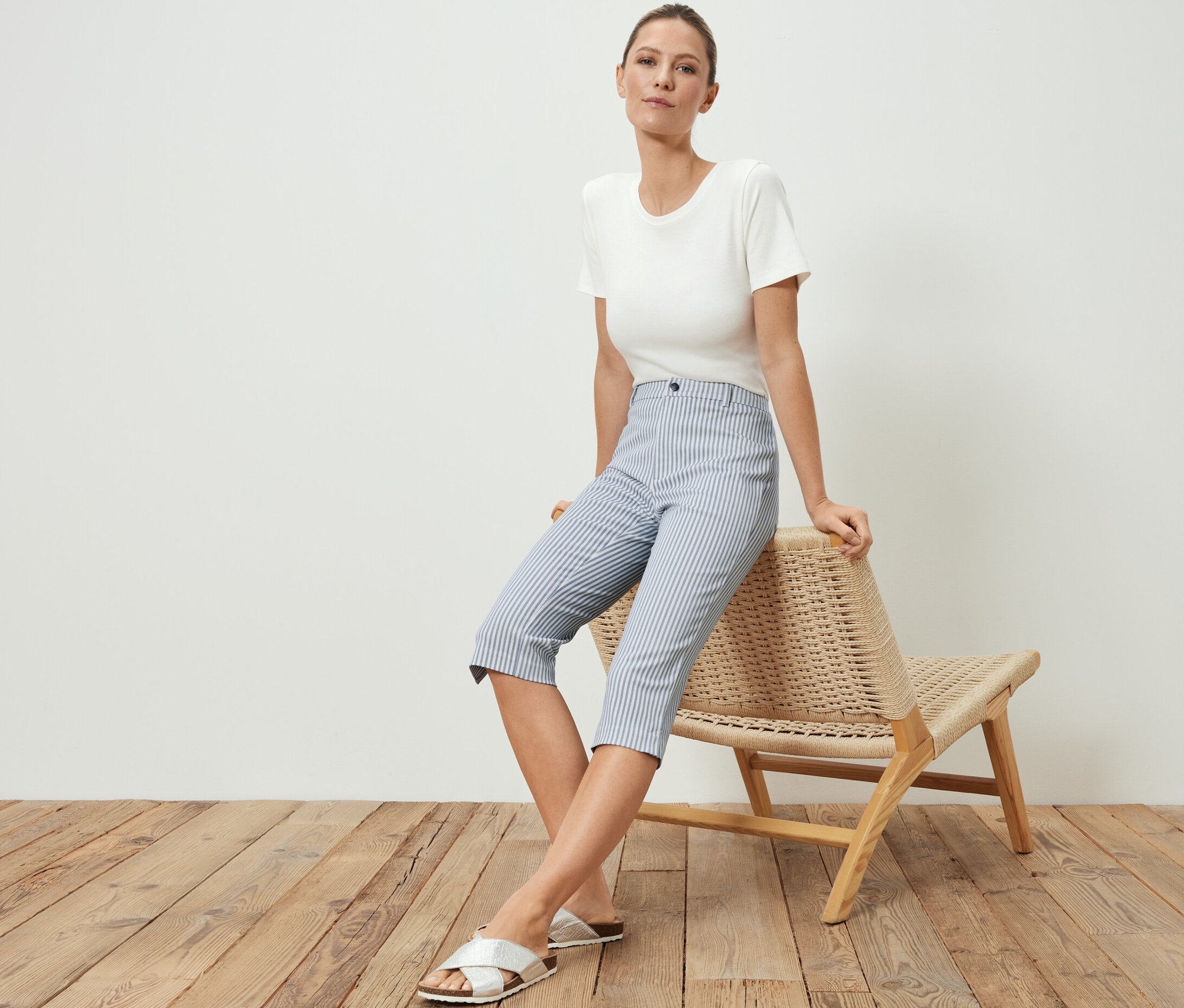 Femme assise sur une chaise en osier portant un pantalon en sergé stretch 3/4, un t-shirt et des sandales.