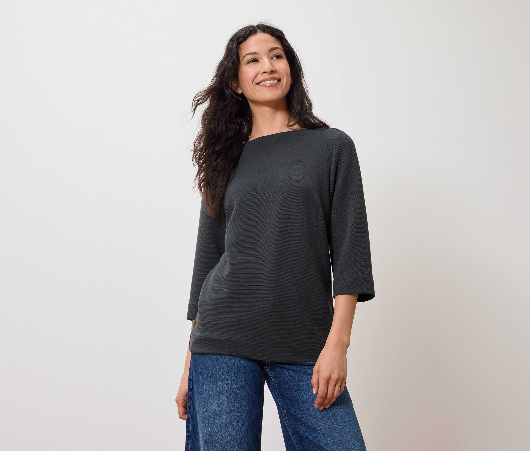 Femme aux cheveux foncés posant dans un sweat-shirt gris à manches trois-quarts et un jean bleu.