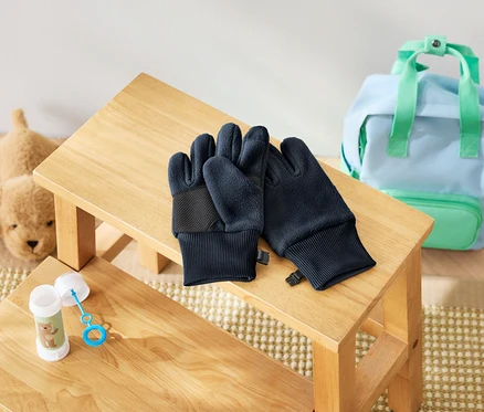 Sur un tabouret en bois, une paire de gants bleu foncé repose à côté d'une bouteille de bulles de savon et d'un chien en peluche. Un sac à dos bleu clair avec des détails vert menthe est visible en arrière-plan.