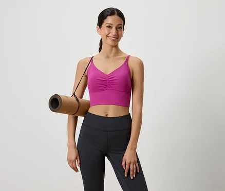 Femme avec un tapis de yoga marron sur l'épaule, vêtue d'un haut de sport rose et d'un legging noir.