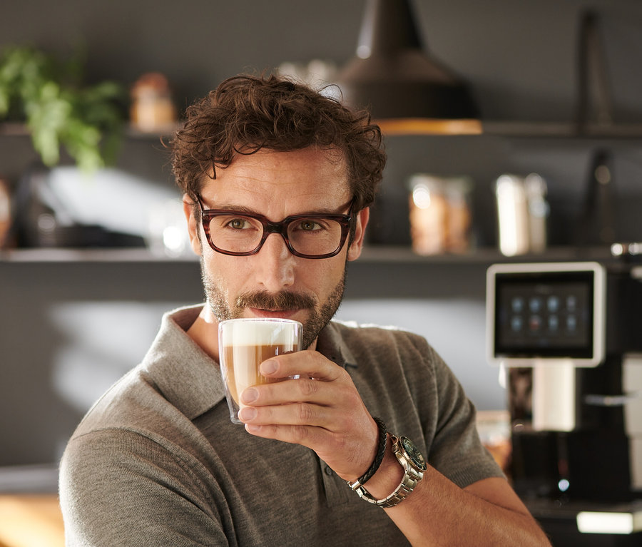 Un homme portant des lunettes boit du café dans un verre transparent. À l'arrière-plan, on voit une cafetière automatique « Tchibo Office », noire.