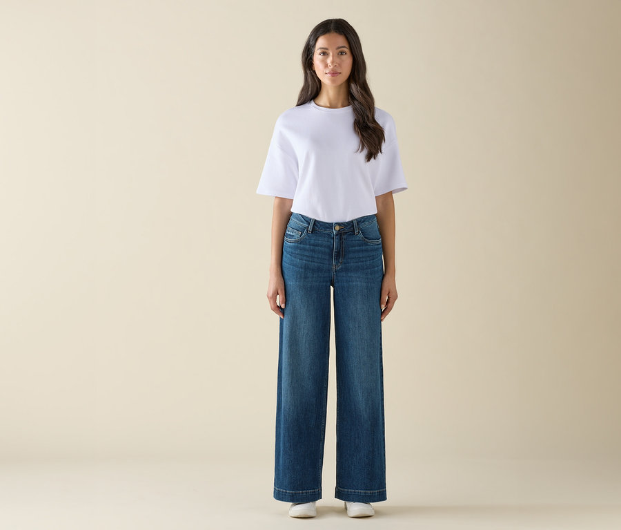 Une femme debout porte un t-shirt blanc, un jean bleu évasé et des baskets blanches.