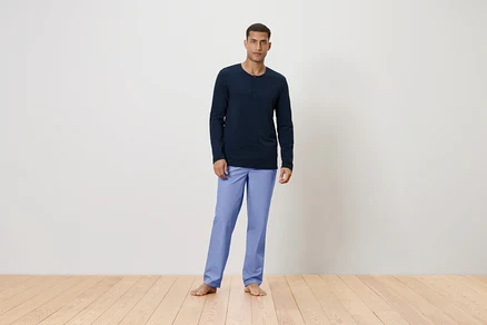 Un homme se tient pieds nus sur un plancher en bois, portant un t-shirt à manches longues bleu marine et un pantalon bleu clair.