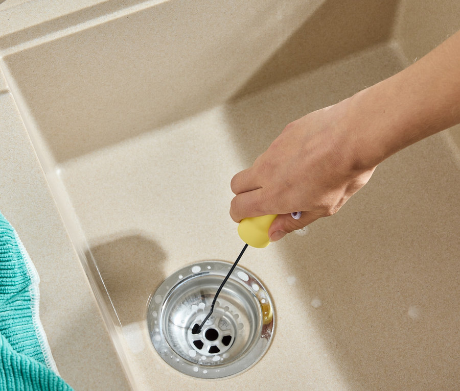 Une main nettoie le drain de l'évier avec un outil jaune. Un torchon vert est à côté de l'évier.