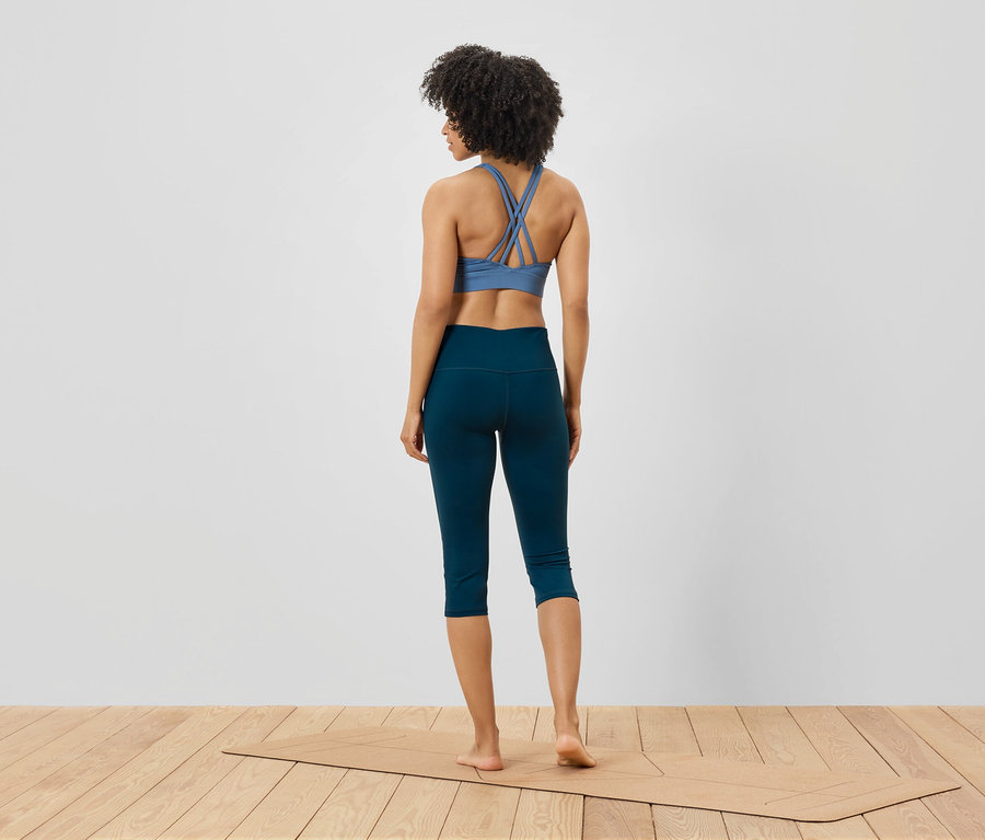 Une femme, vue de dos, porte un soutien-gorge de sport bleu et un legging bleu marine, et se tient sur un tapis de yoga.