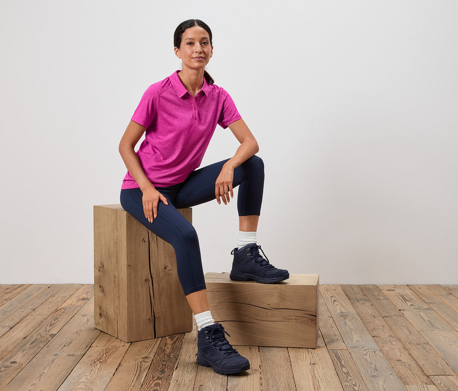 Une femme est assise sur un tabouret en bois, portant un polo rose, un legging bleu et des chaussures de randonnée bleues.