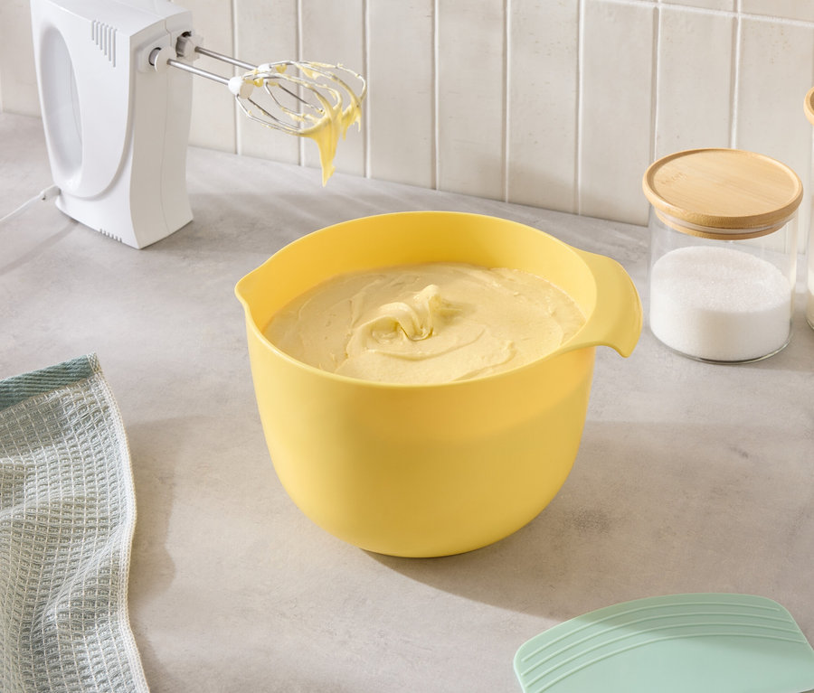 Sur le comptoir de la cuisine, on voit un bol jaune rempli de pâte, un mixeur et des bocaux de sucre et de farine.
