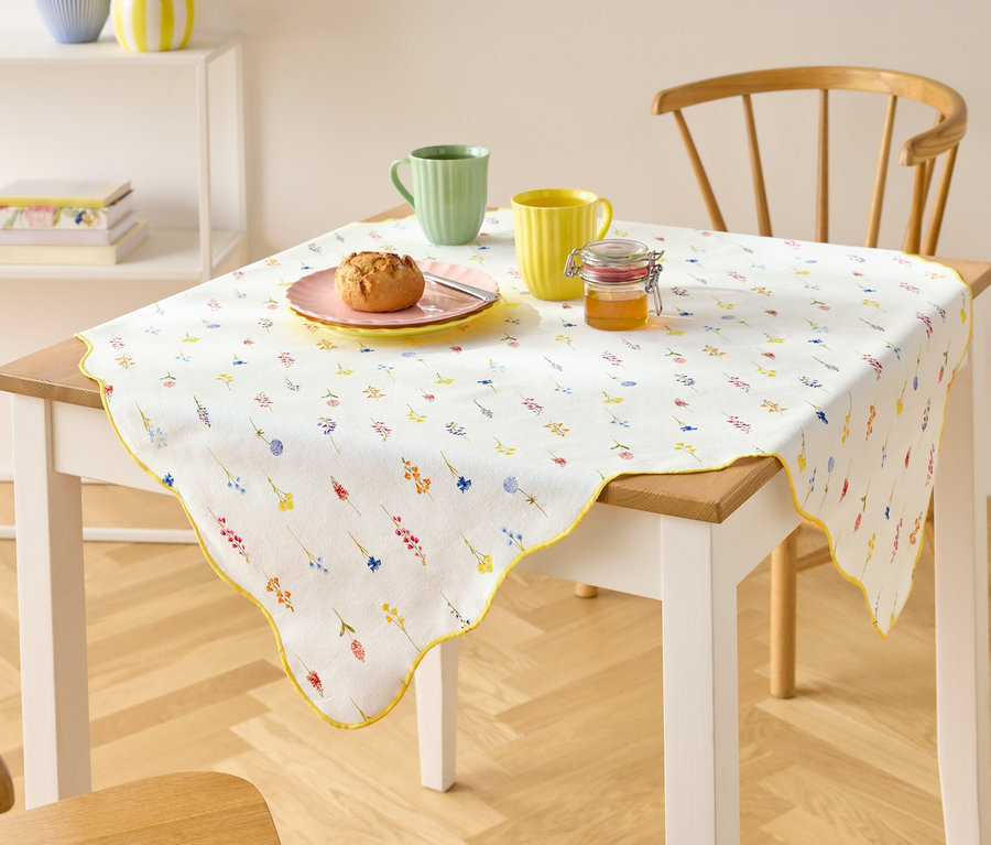 Une table avec une nappe blanche à motifs floraux, deux tasses, une assiette avec un pain et un pot de miel.