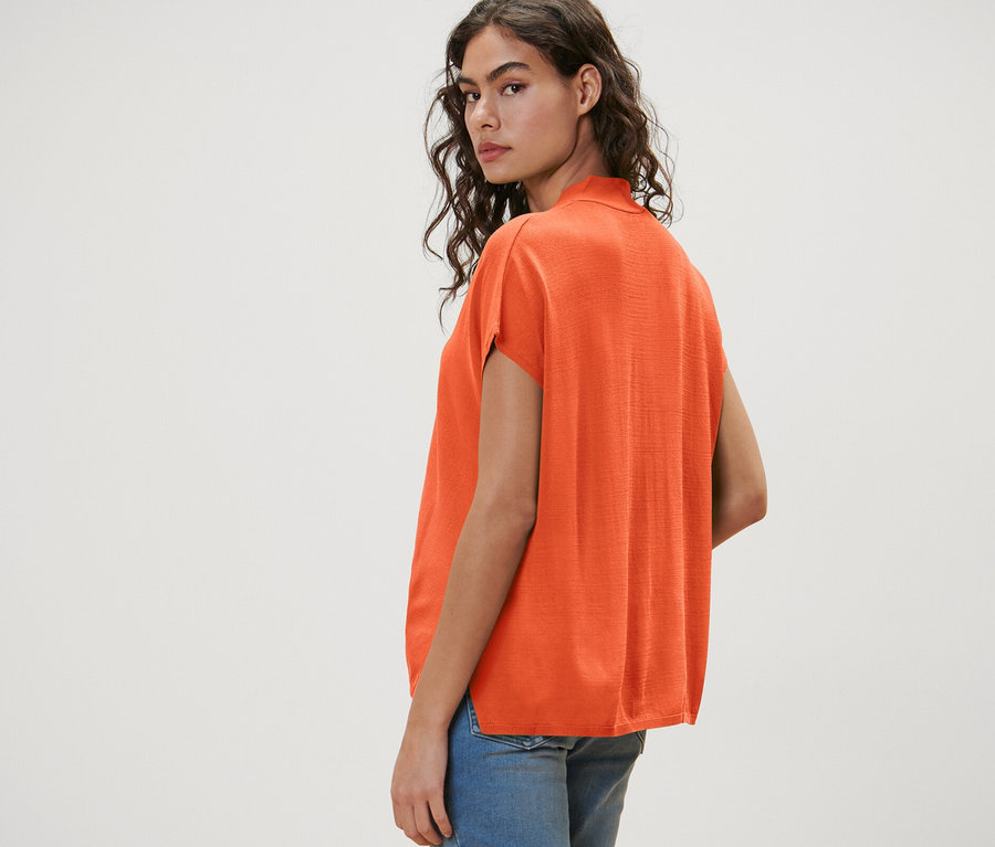 Une femme regarde par-dessus son épaule et porte un cardigan orange à manches courtes et un short en jean.