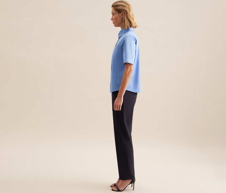 Femme de profil, portant un chemisier à manches courtes Seidensticker bleu clair, un pantalon foncé et des sandales noires.