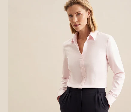 Une femme aux cheveux blonds porte un chemisier en tulle Seidensticker rose et un pantalon foncé, les mains dans les poches.