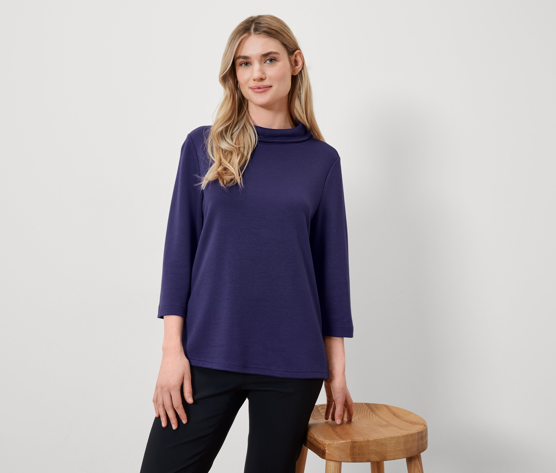 Une femme blonde pose dans un haut bleu marine à manches trois-quarts et un pantalon noir à côté d'un tabouret en bois.