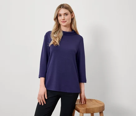 Une femme blonde pose dans un haut bleu marine à manches trois-quarts et un pantalon noir à côté d'un tabouret en bois.