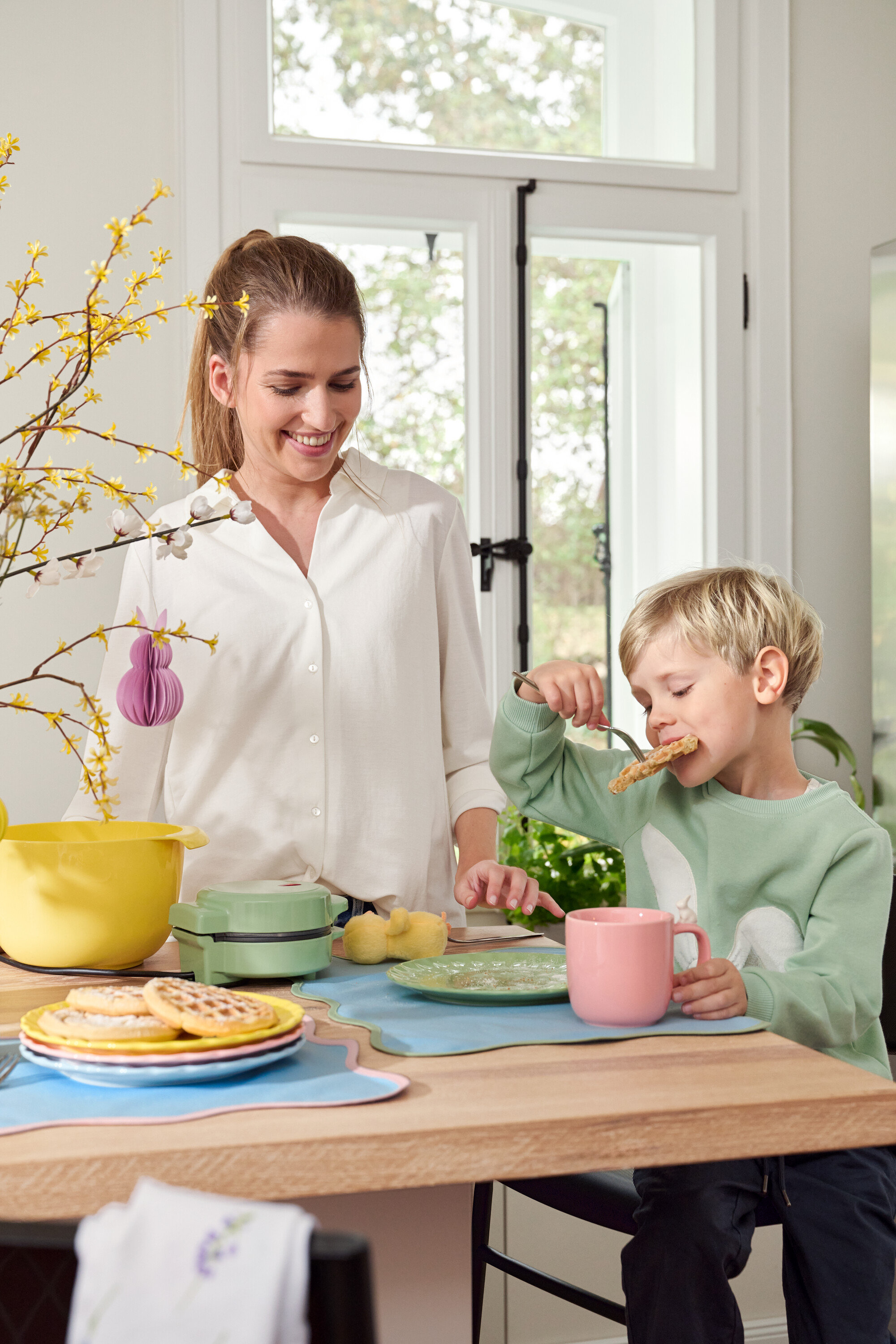 Femme et enfant à table. L'enfant mange une gaufre avec une fourchette, la femme sourit.