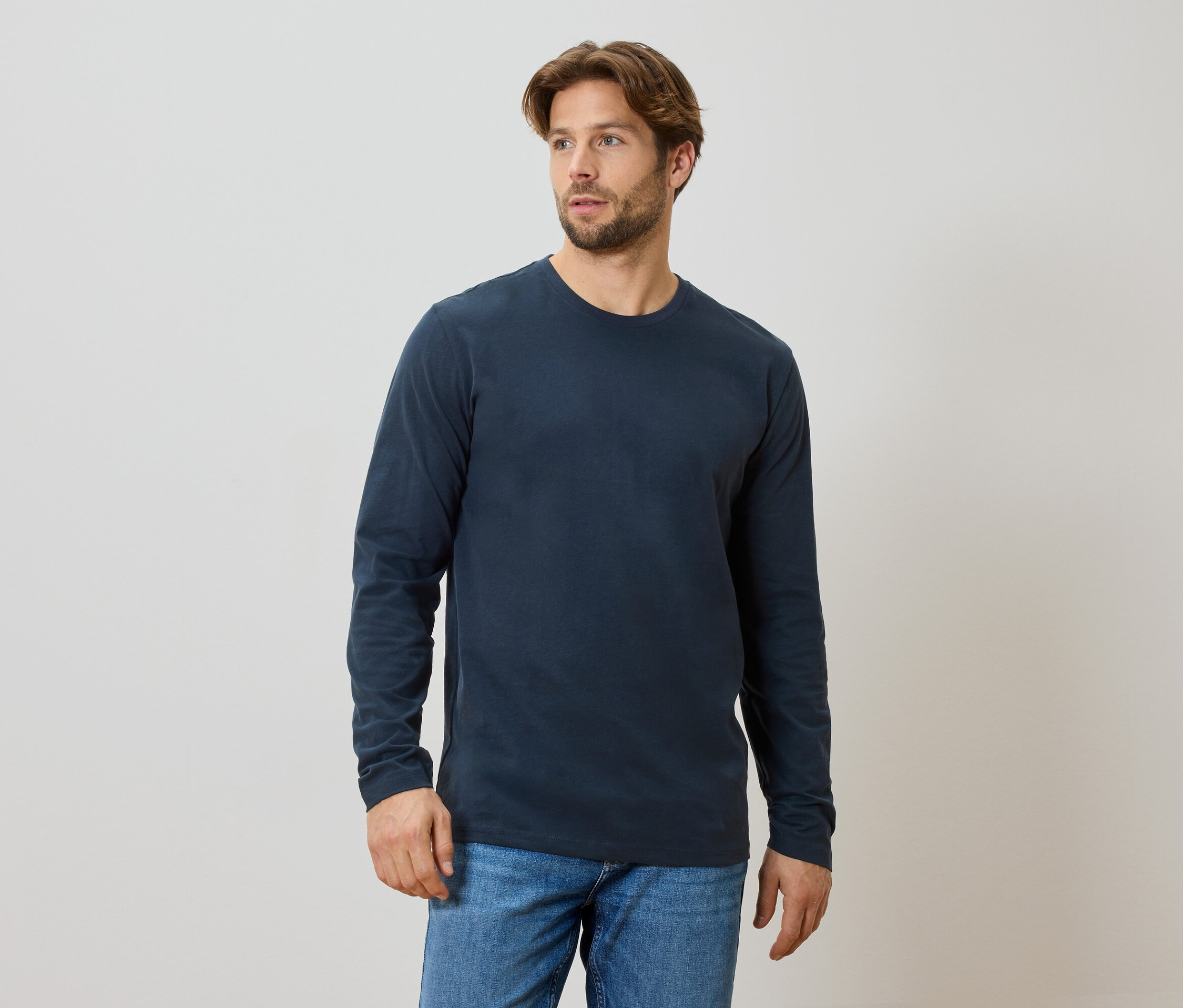 Un homme barbu porte un t-shirt bleu marine à manches longues et un jean.