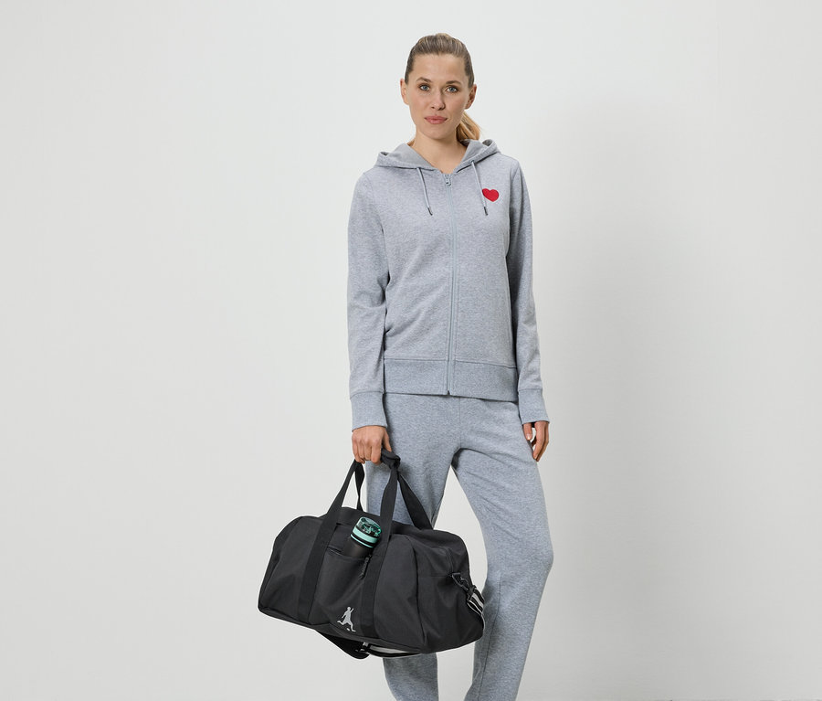 Femme portant un survêtement gris avec un cœur rouge et tenant un sac de sport noir.