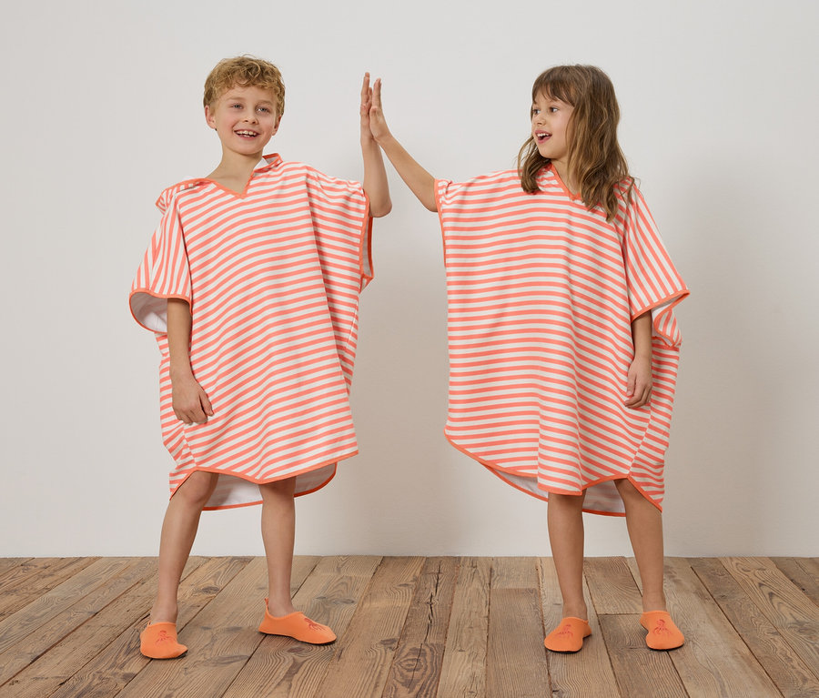 Deux enfants, un garçon et une fille, se tiennent sur un plancher en bois devant un mur blanc et se font un high-five. Ils portent tous les deux des ponchos rayés orange et blanc et des chaussures oranges.