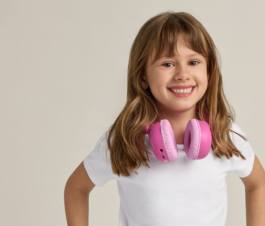 Fillette souriante avec de longs cheveux bruns et un casque rose autour du cou, sur un fond clair.