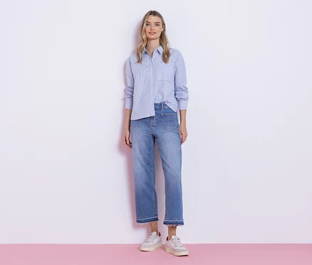 Une jeune femme se tient devant un mur blanc, vêtue d'un chemisier Street One rayé bleu et blanc, d'un jean clair à ourlet effiloché et de baskets blanches.