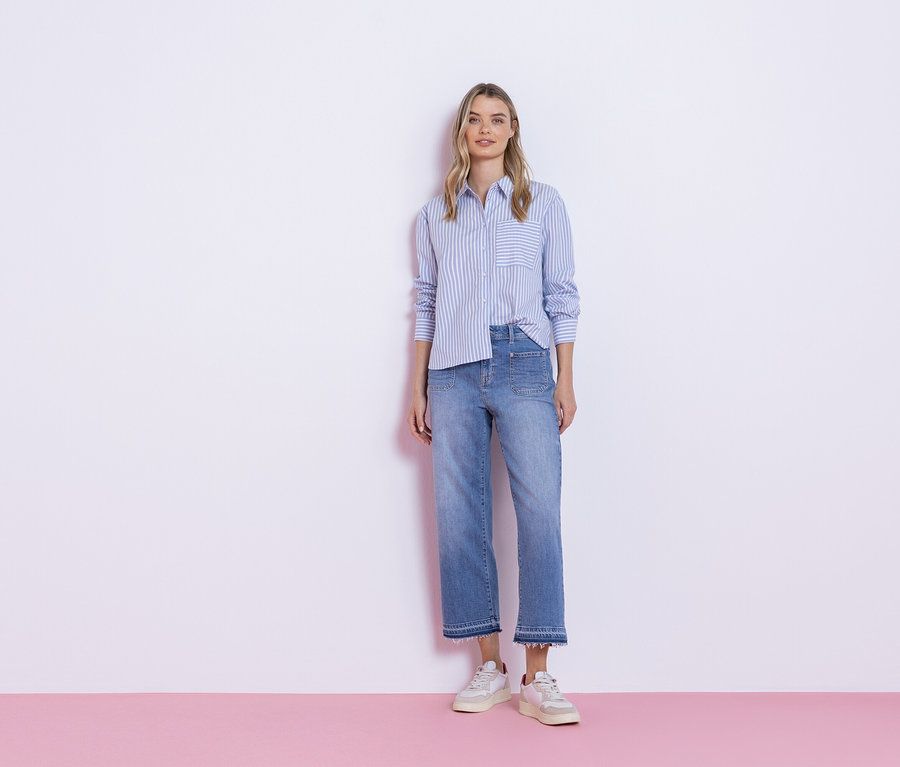 Une femme se tient devant un mur blanc, vêtue d'une chemise à rayures bleues, d'un jean et de baskets.