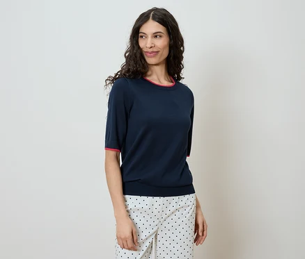 Une femme aux cheveux ondulés et foncés porte un T-shirt bleu marine à manches courtes avec une bordure rouge et un pantalon à pois sur un fond clair.