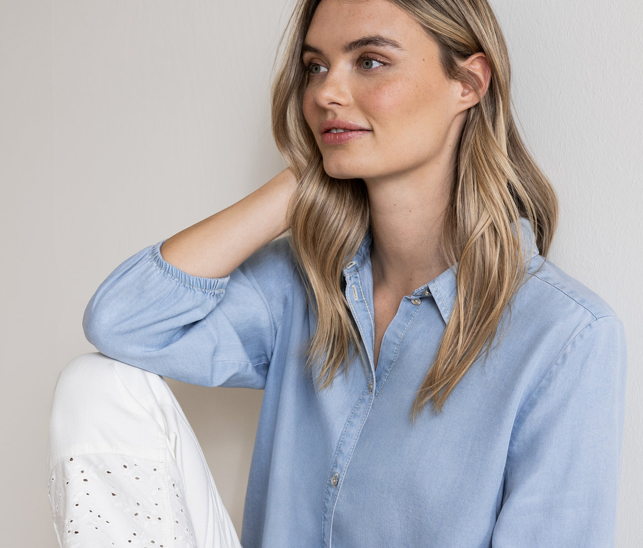 Une femme blonde porte une chemise bleu clair et un pantalon blanc ajouré.