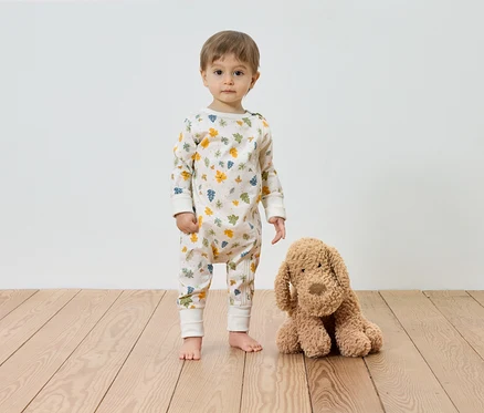 Un enfant se tient pieds nus à côté d'un chien en peluche sur un plancher en bois devant un mur blanc. L'enfant porte une combinaison à motifs de feuilles.