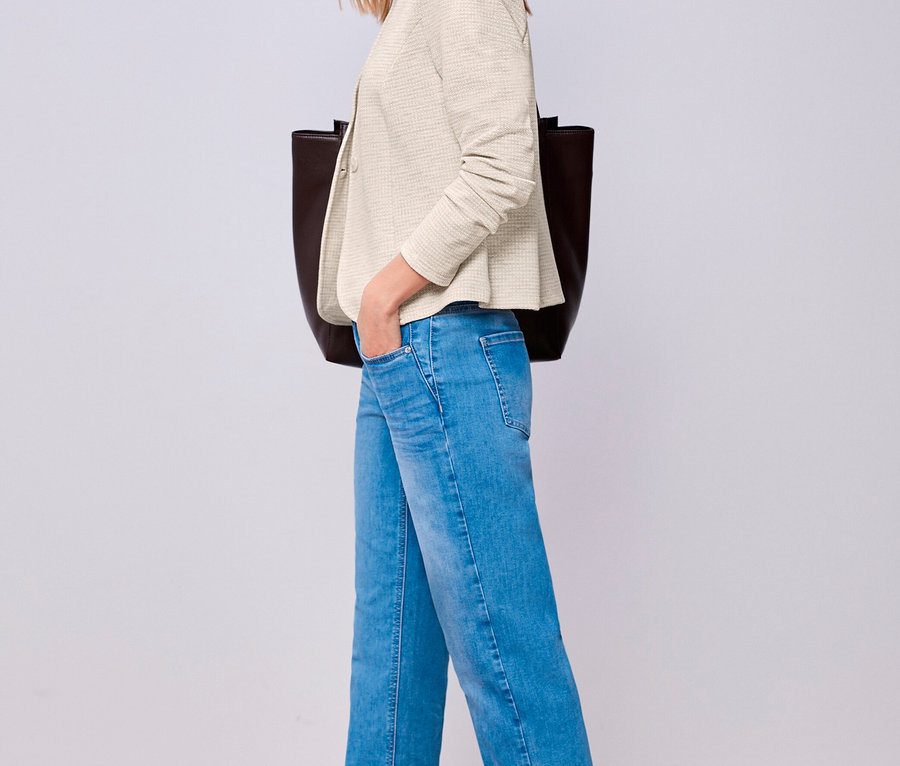 Femme portant un blazer en jersey Street One crème, un jean bleu et des chaussures plates foncées, une main dans la poche, avec un sac cabas foncé.