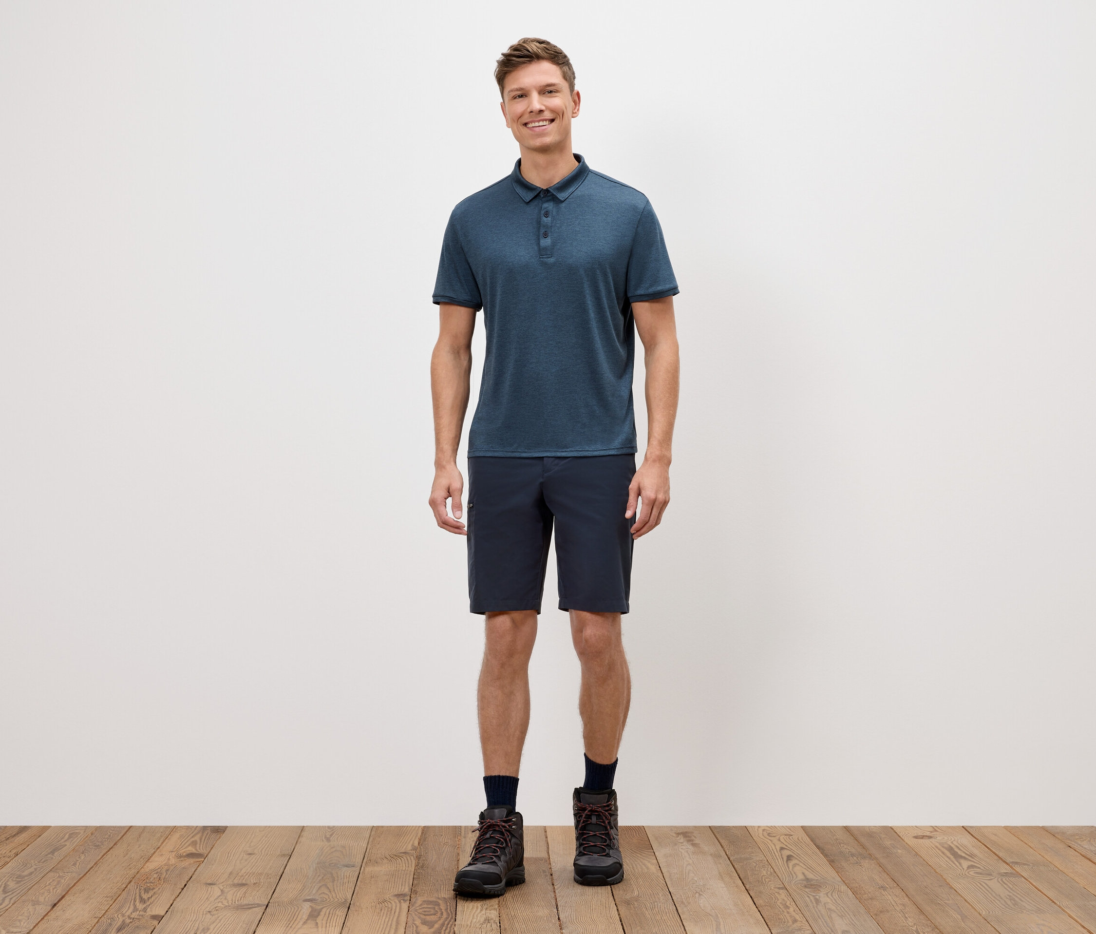 Un homme se tient devant un mur blanc, portant un polo bleu, un short et des chaussures de randonnée.