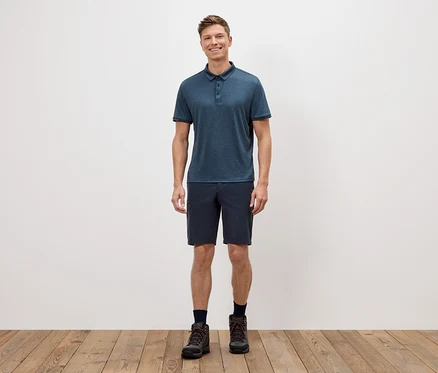 Un homme se tient devant un mur blanc, portant un polo bleu, un short et des chaussures de randonnée.