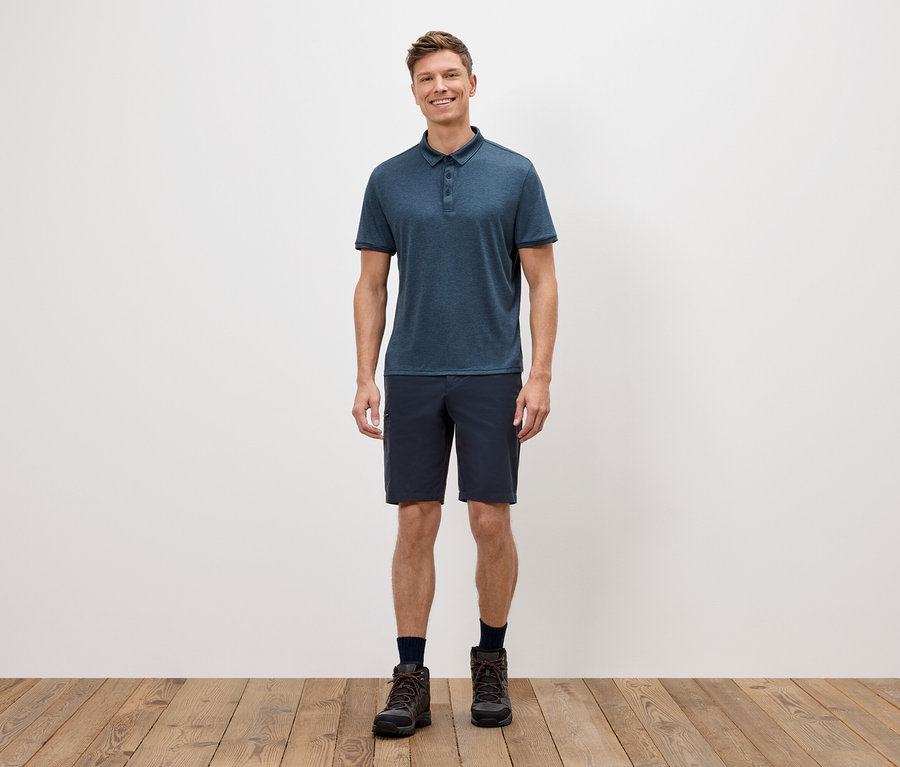 Un homme se tient devant un mur blanc, portant un polo bleu, un short et des chaussures de randonnée.