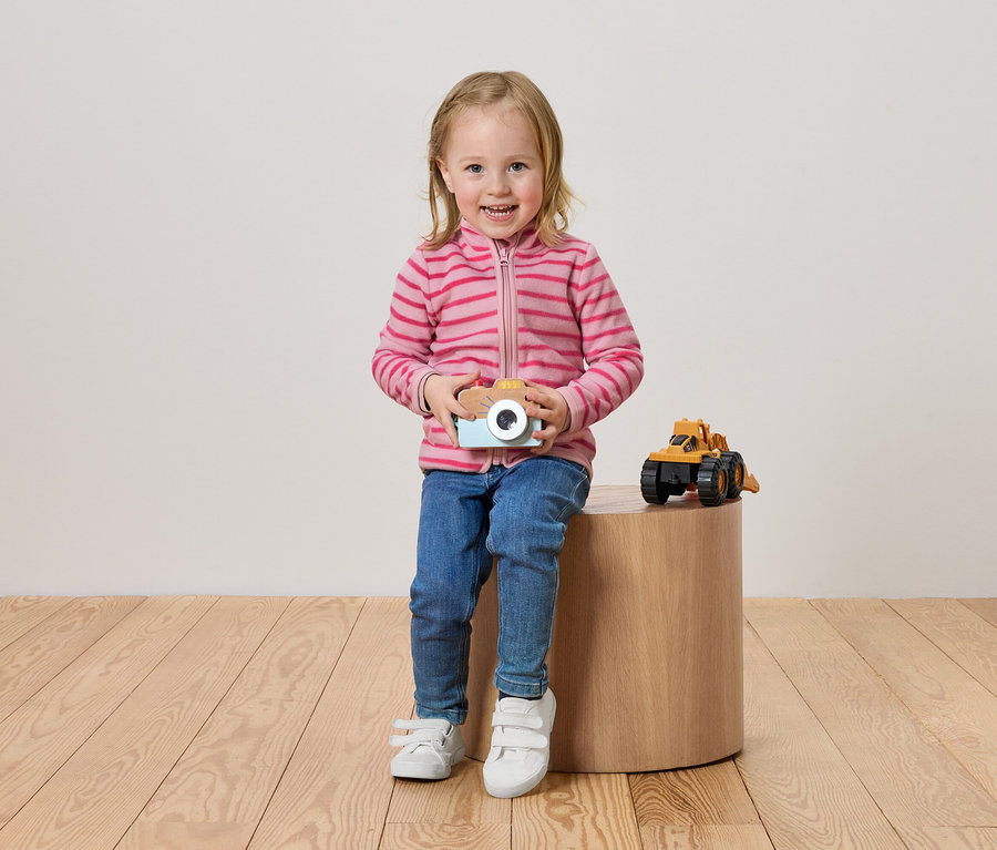 Une petite fille est assise sur un tabouret en bois et tient un appareil photo jouet dans ses mains. À côté d'elle se trouve une pelleteuse jouet jaune.