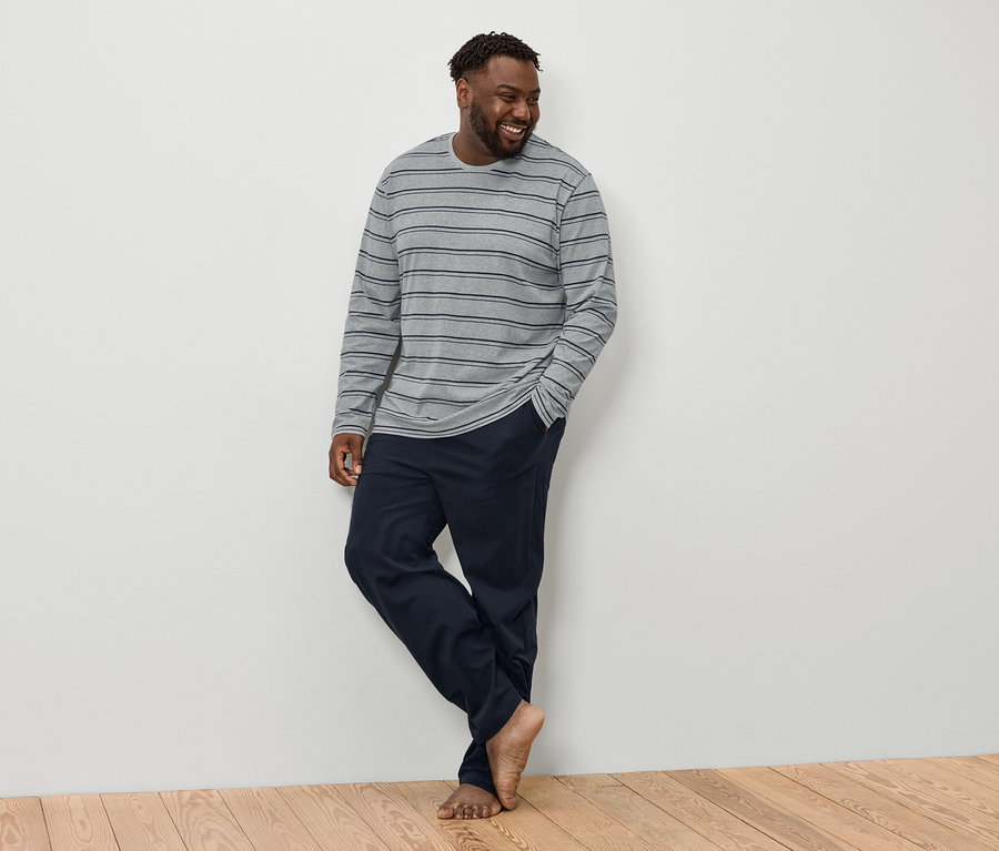 Un homme souriant aux cheveux foncés et à la barbe se tient pieds nus, vêtu d'un T-shirt à manches longues gris rayé et d'un pantalon bleu marine.