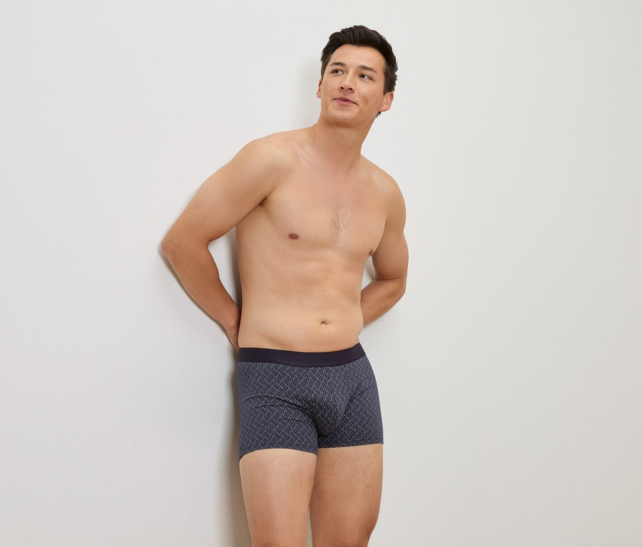 Homme torse nu appuyé contre un mur blanc portant un boxer bleu à motifs.