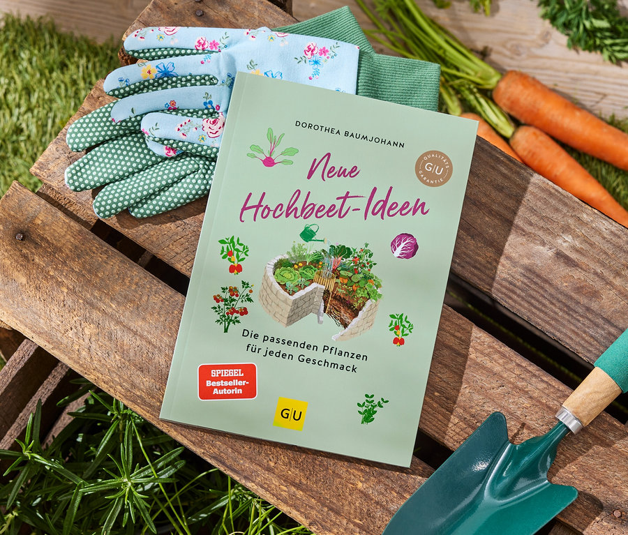 Des gants de jardinage, des carottes, une pelle de jardin verte et le livre 'Neue Hochbeet-Ideen' sont posés sur une caisse en bois.