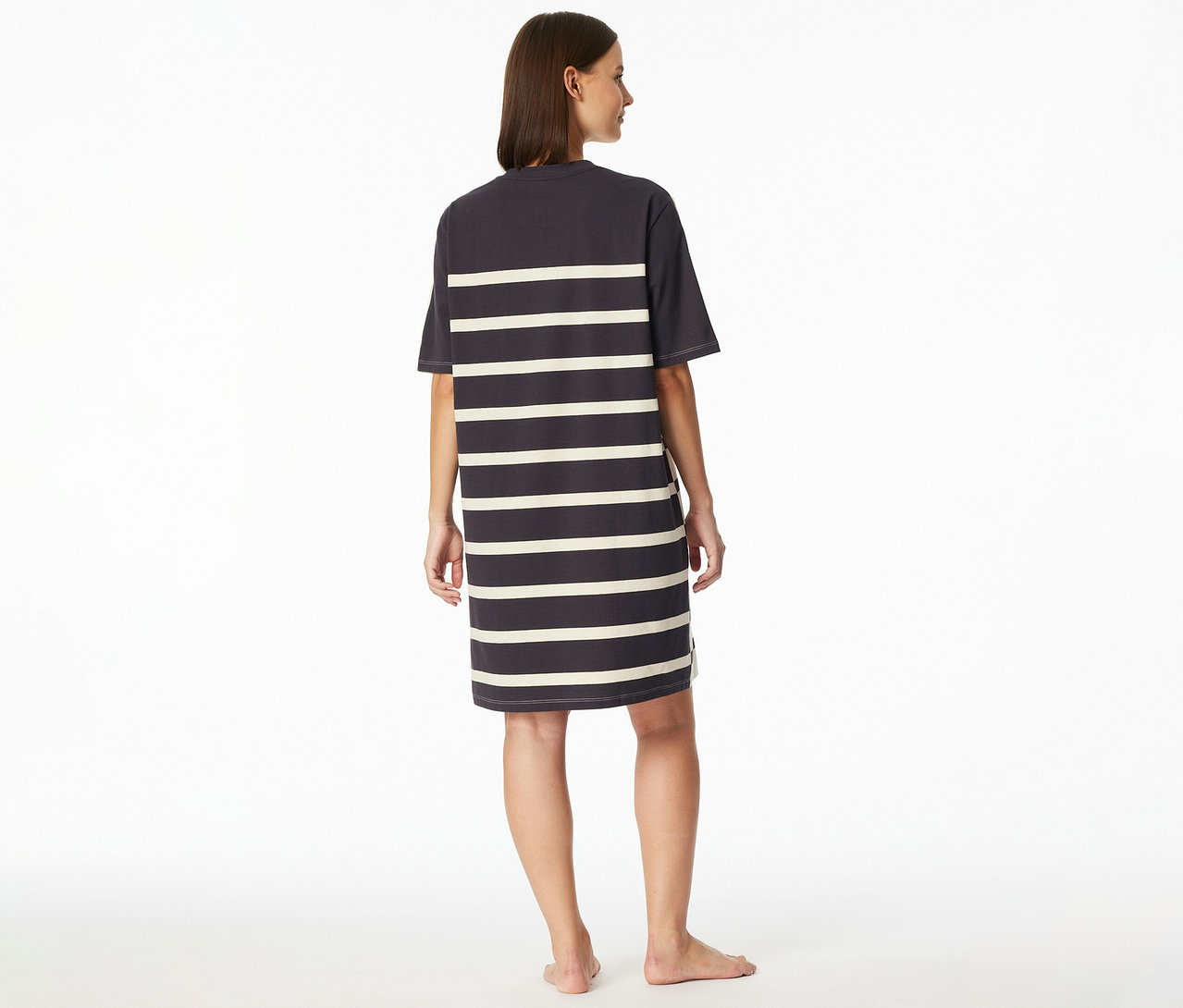 Une femme est debout pieds nus, dos à la caméra, regardant légèrement à droite, vêtue d'une chemise de nuit SCHIESSER rayée.