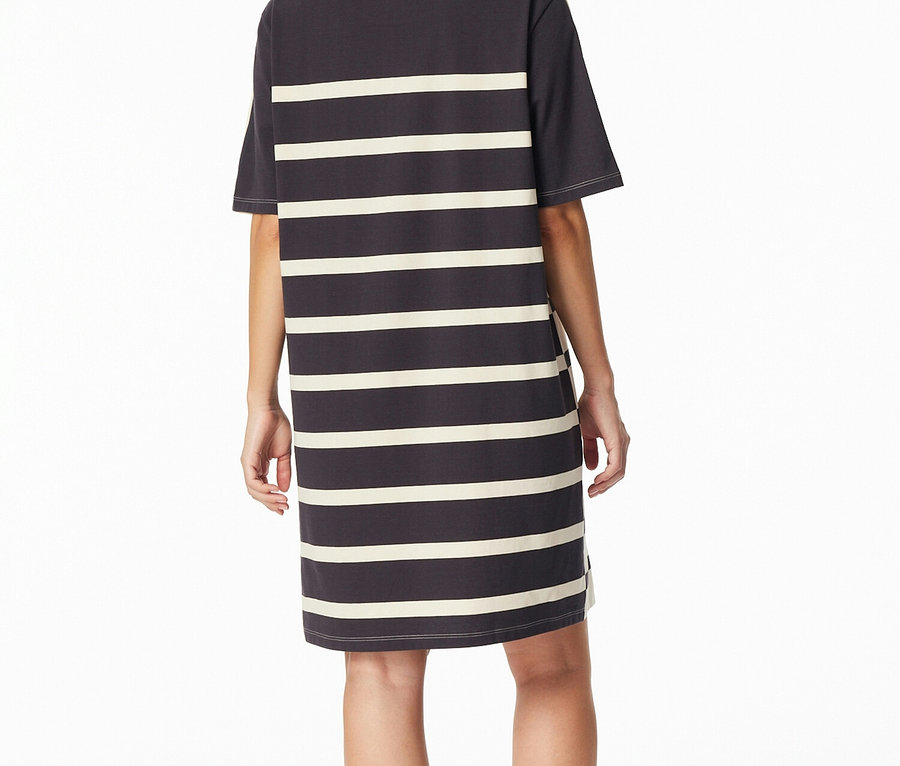 Une femme est debout de dos, vêtue d'une chemise de nuit SCHIESSER gris foncé à rayures horizontales crème.