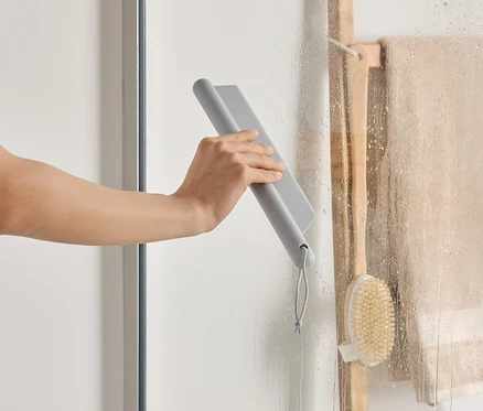 Une main essuie une paroi de douche avec une raclette grise, à côté d'une échelle en bois avec une serviette et une brosse.