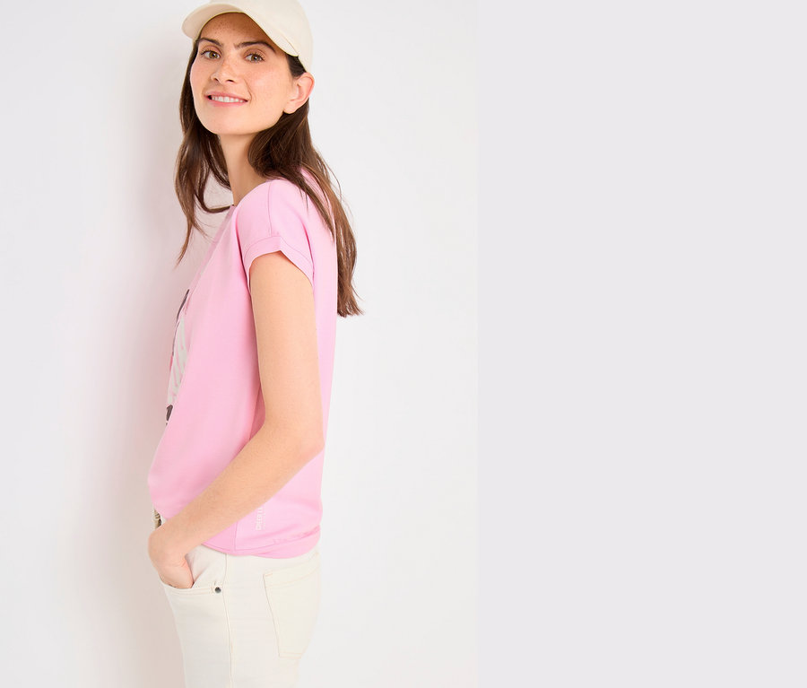 Femme souriante portant une casquette claire et un pantalon clair, vêtue d'un T-shirt à motif CECIL rose aux manches retroussées, mains dans les poches.