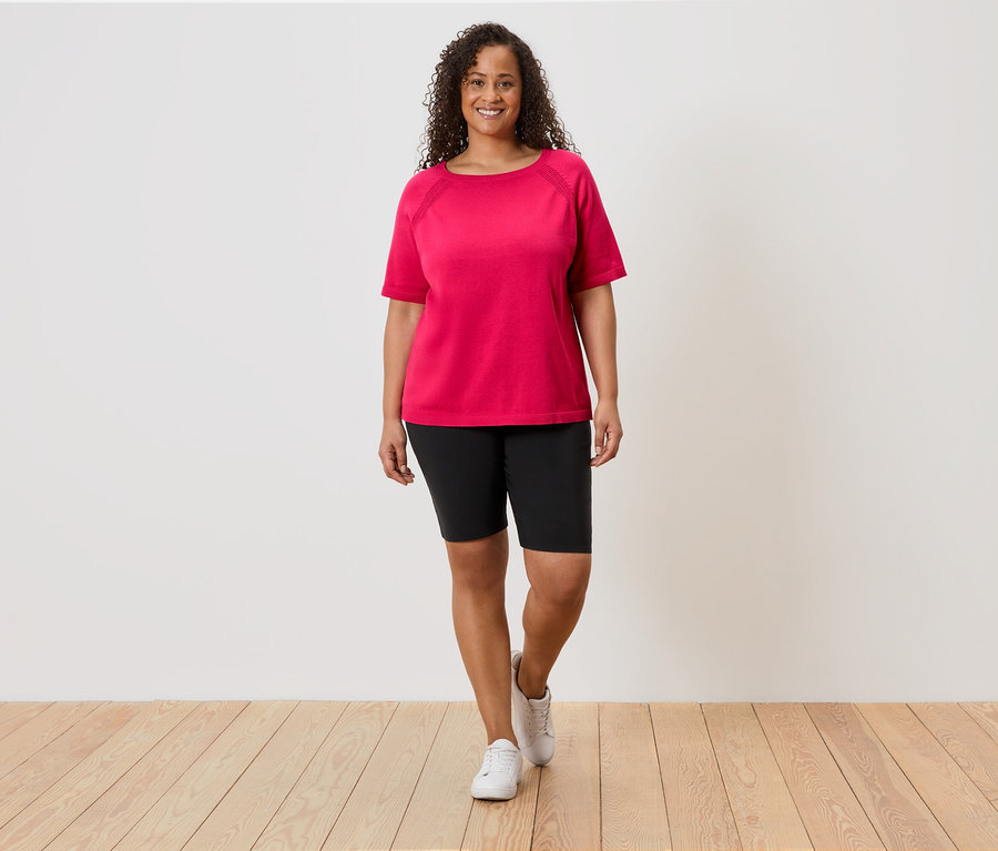 Une femme aux cheveux bouclés porte un t-shirt rose, un short noir et des baskets blanches devant un mur blanc.