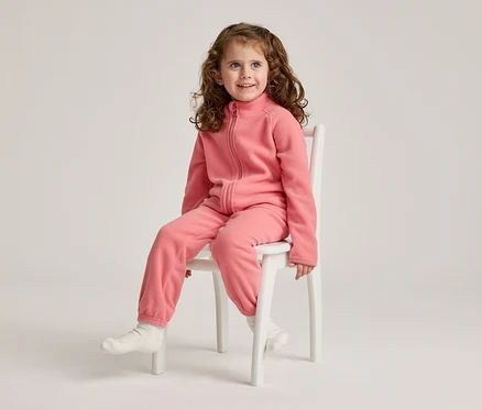 Une fillette aux cheveux bouclés est assise sur une chaise blanche et porte une combinaison en polaire rose et des chaussettes blanches.