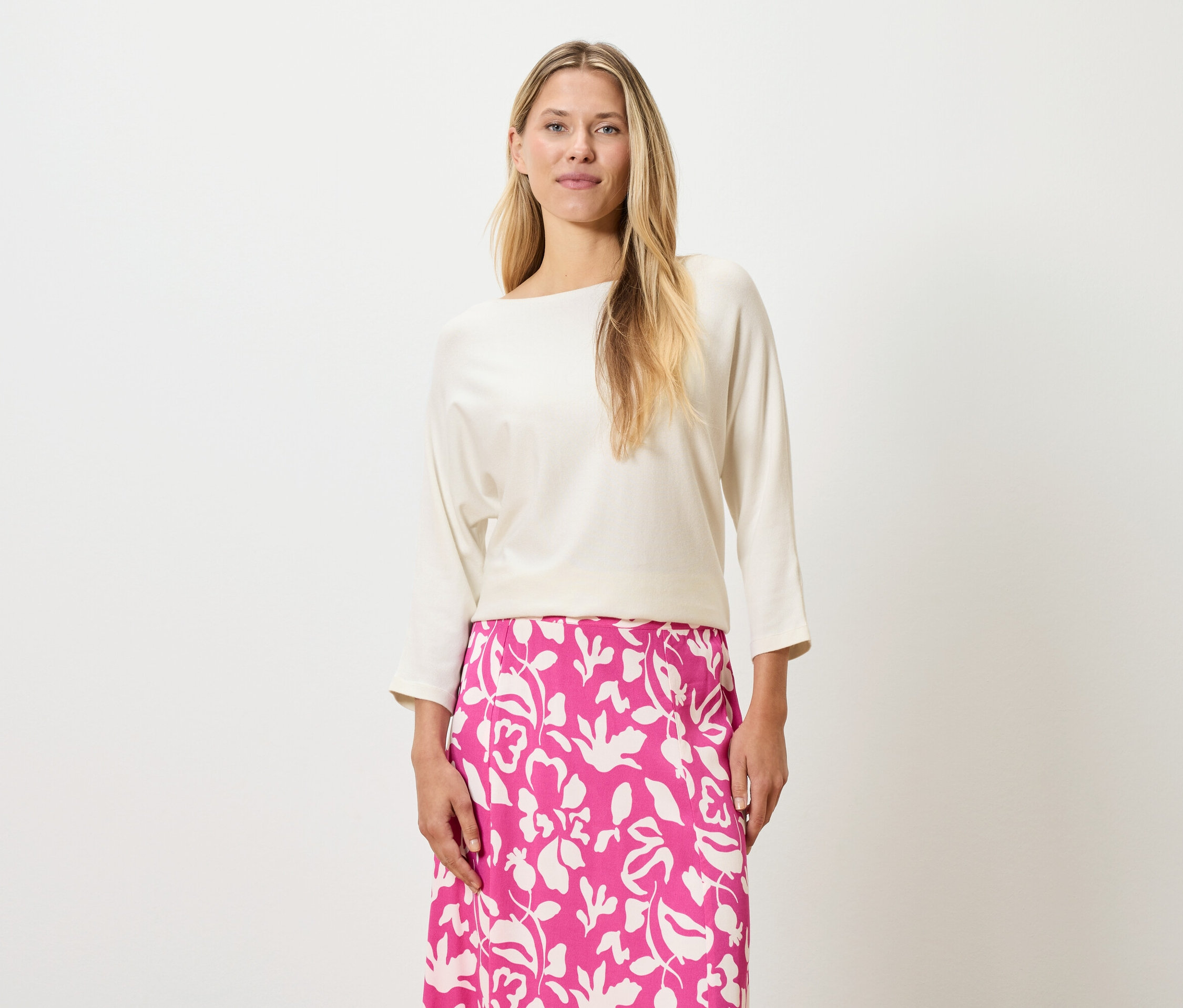 Une femme aux cheveux blonds porte un haut couleur crème et une jupe rose à imprimé floral.
