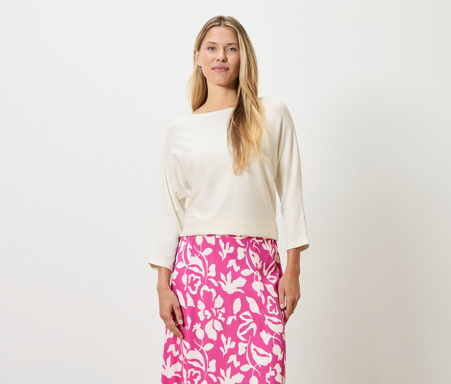 Une femme aux cheveux blonds porte un haut couleur crème et une jupe rose à imprimé floral.