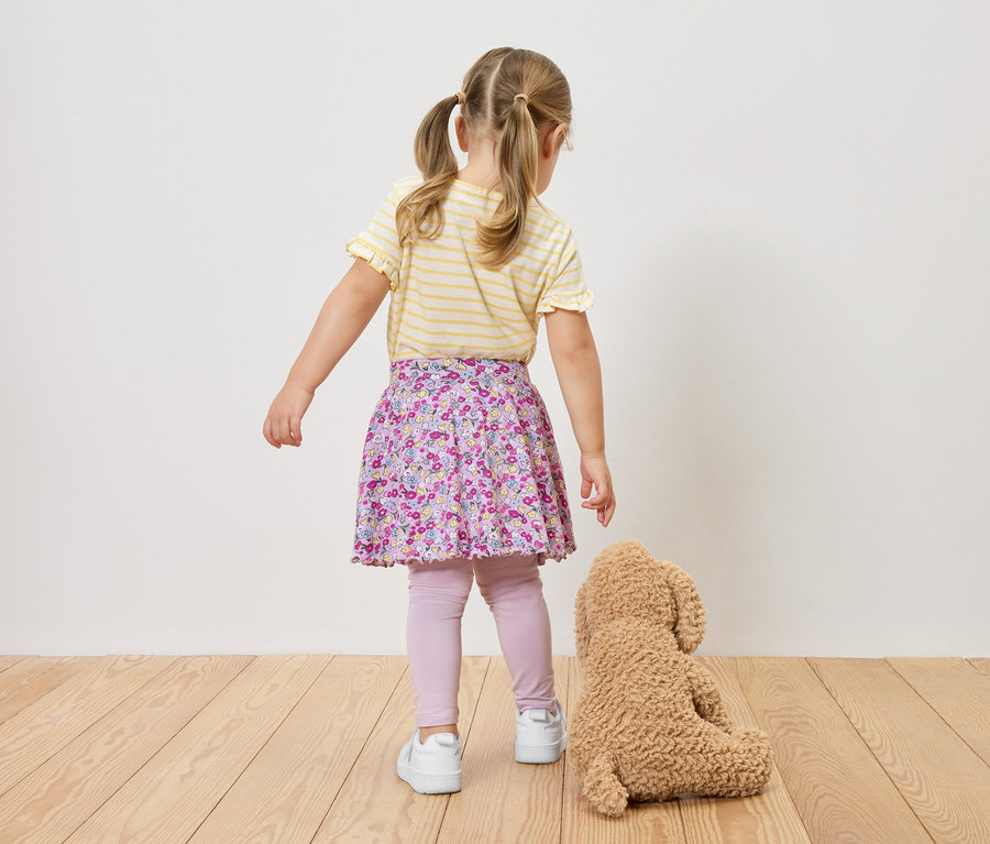 Vue de dos d'une fillette avec des tresses portant un haut rayé jaune et blanc, une jupe à fleurs, des leggings et des baskets, à côté d'un ours en peluche.